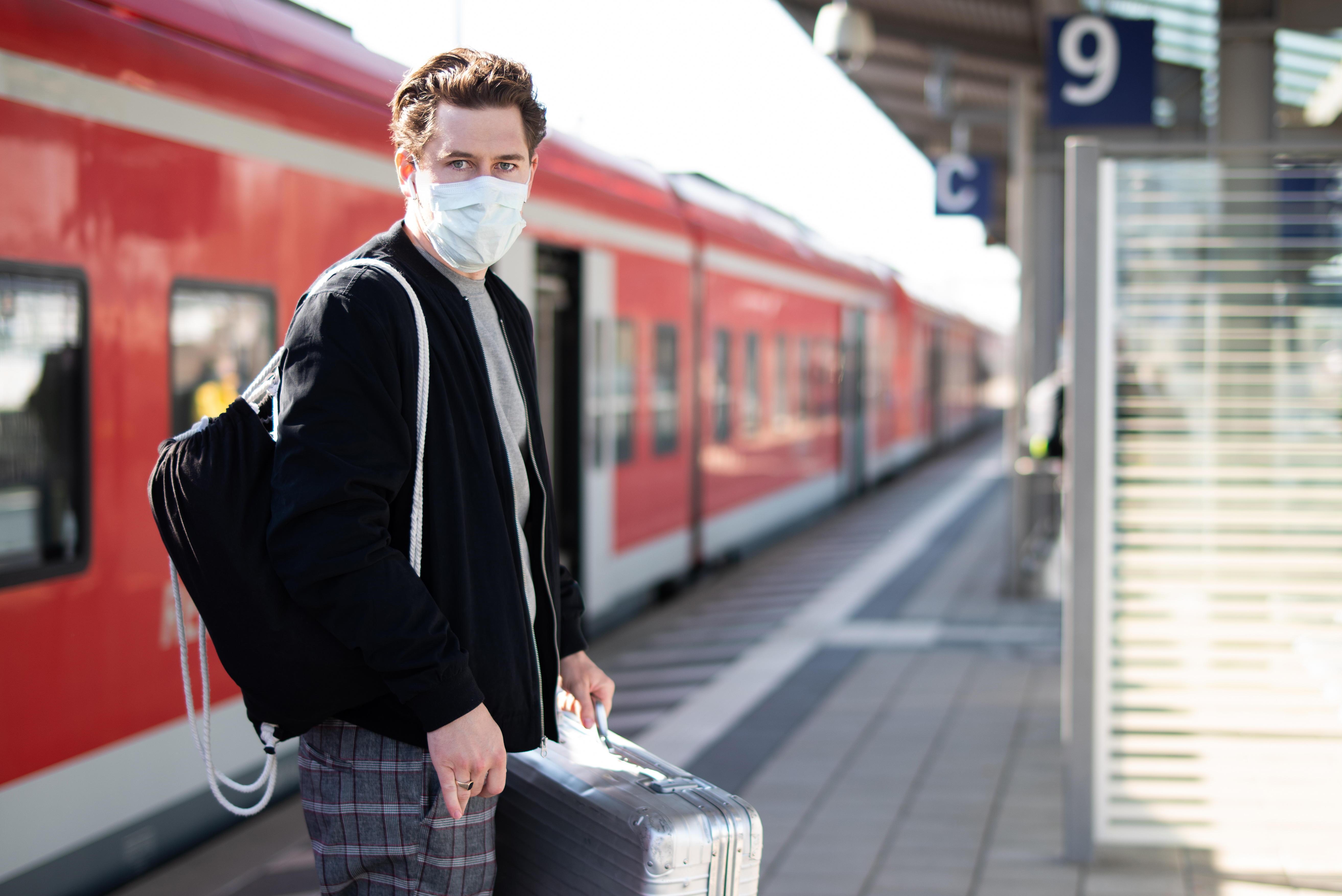 Mann mit Koffer auf dem Bahnsteig, er schützt sich mit Mundschutz und Handschuhen vor einer Ansteckung mit dem Coronavirus