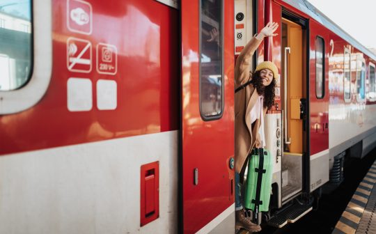 Junge Frau glücklich mit Koffer winkt aus Tür im Regionalzug