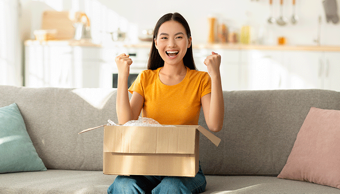 Umgang mit NPS-Feedback - junge Frau öffnet ein Paket und zeigt ihre Begeisterung über einen erfolgreichen Produktkauf indem sie lacht und ihre Hände in die Luft streckt