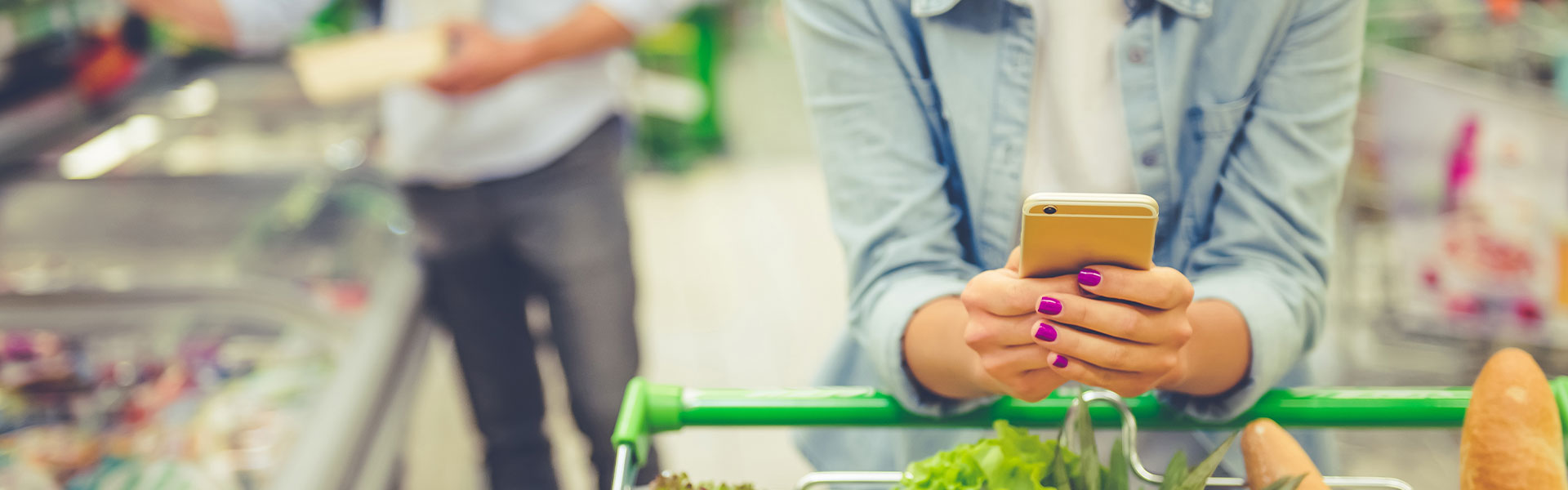 Mit dem NPS Kundenzufriedenheit ermitteln. Eine Frau ist im Supermarkt mit einem Einkaufswagen voller Gemüse und hat ihr Handy in der Hand.