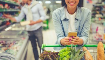 Mit dem NPS Kundenzufriedenheit ermitteln. Eine Frau ist im Supermarkt mit einem Einkaufswagen voller Gemüse und hat ihr Handy in der Hand.