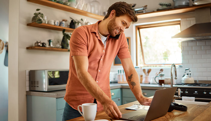 Chancengleichheit erfolgreich umsetzen: Ein Mann steht in der Küche mit einer Tasse Kaffee, Laptop und seinem Handy am Ohr, um zu arbeiten.